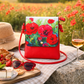 Red handbag with floral poppy design on a picnic setting with a glass of wine, food, and sunglasses.